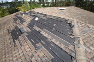 Roof of a house showing extensive shingle damage, many missing/curling tiles, and exposed black underlayment with debris; palm trees in the background.