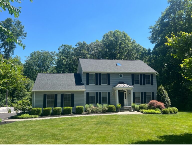 rogan1 - Custom Home Renovations Two-story gray suburban house with black shutters and a small front porch, surrounded by a manicured lawn and shrubs under a clear blue sky.